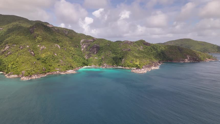 A lush green island with rocky cliffs and a small turquoise bay is surrounded by clear blue ocean under a partly cloudy sky