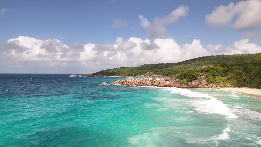 Flying low over white sand as waves come ashore the camera rises higher to reveal the stunning turquoise water and a rugged jagged coastline
