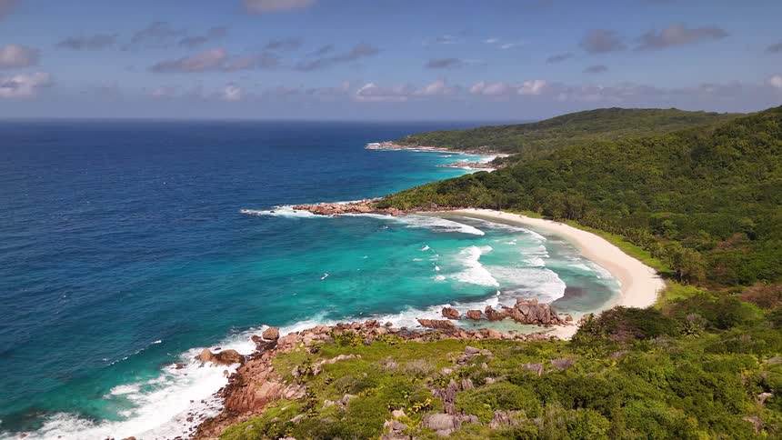A sandy beach curves beside turquoise waters and rocky outcrops framed by lush hills under a partly cloudy sky
