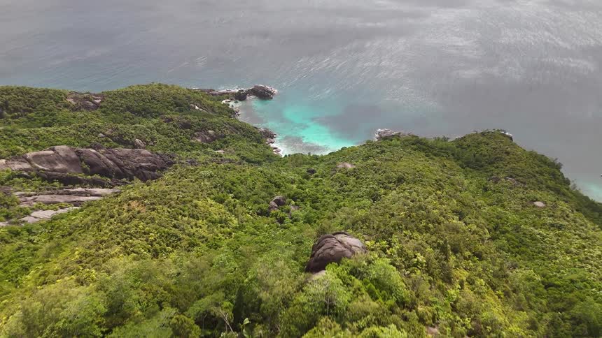 A green mountain ridge with scattered boulders overlooks crashing turquoise waves along a rugged treecovered coastline