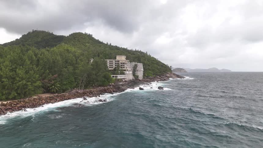 A building sits on a rocky coastline surrounded by lush green hills and turbulent ocean waves under a cloudy sky