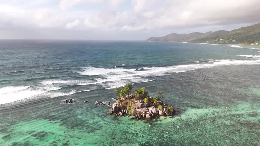 A small rocky island sits in the middle of the ocean with waves crashing onto it at midday