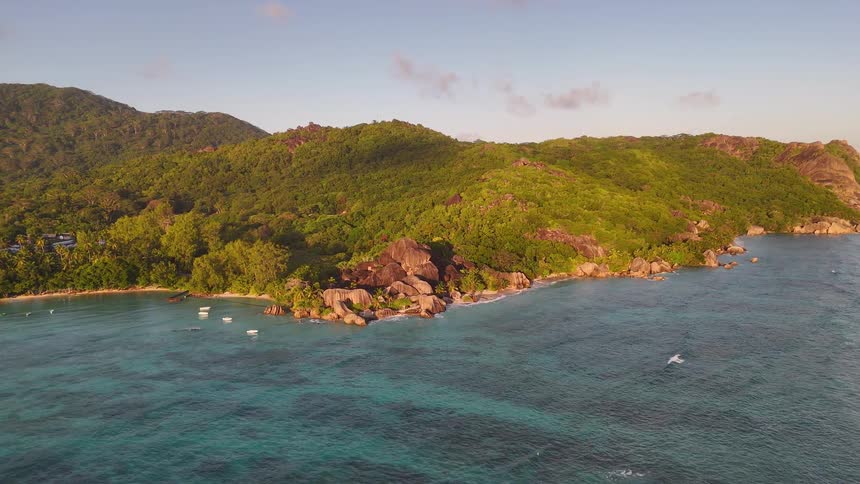 An aerial shot captures a rocky tropical coastline during golden hour showing small boats moored in shallow water as gentle waves come ashore