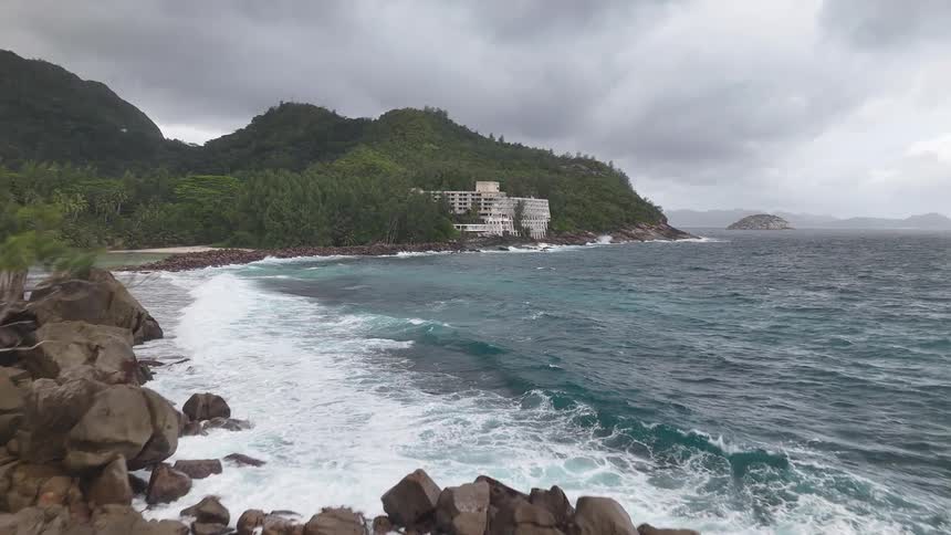 A camera rapidly flies forward in an aerial view moving over a rocky ocean jetty and toward a large hotel on a tropical coastline