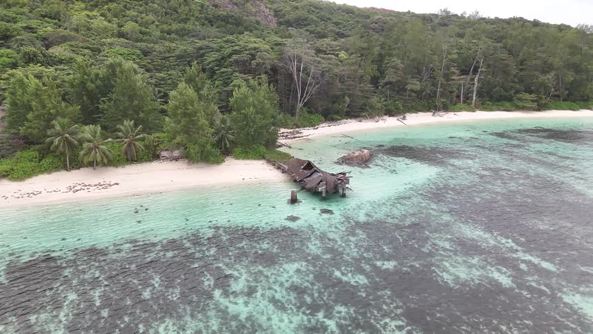 An aerial shot flies sideways over a tropical island showing a destroyed pier and ruined building on a white sand beach by clear shallow water