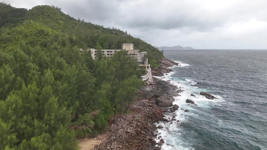 A cliffside building stands against crashing waves and lush forested hills under a cloudy sky