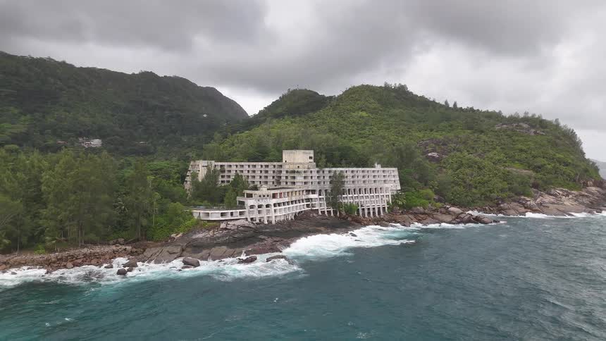 A large multistory building stands on a rocky shoreline with waves crashing below surrounded by dense green hills under a cloudy sky