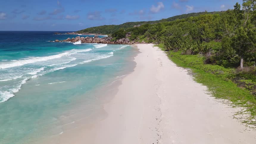 An aerial view flies slowly forward along a deserted tropical beach with white sand turquoise water and a lush green forest