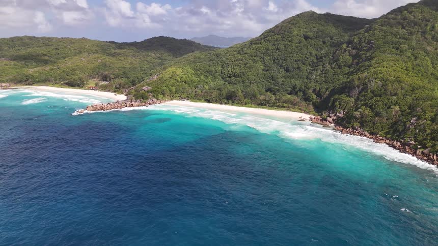 A stunning aerial view pulls backward and away from a tropical coastline showing a secluded white sand beach nestled between the ocean and lush mountains
