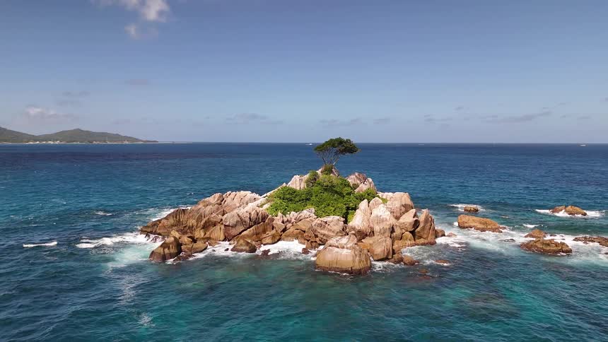A stunning aerial shot circles a small rocky islet that features a solitary tree revealing turquoise water and distant islands in the background
