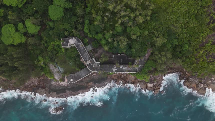 An aerial view flies upwards from a derelict building on an island showing waves coming ashore on the rocky coast next to a dense forest