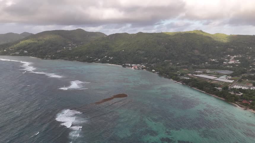 A coastal town stretches along coral shallows nestled beneath lush green mountains and a partly cloudy sky