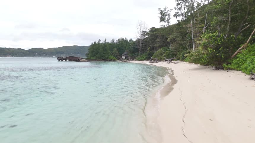 Clear turquoise water surrounds a treelined beach with a distant weathered dock jutting into the bay