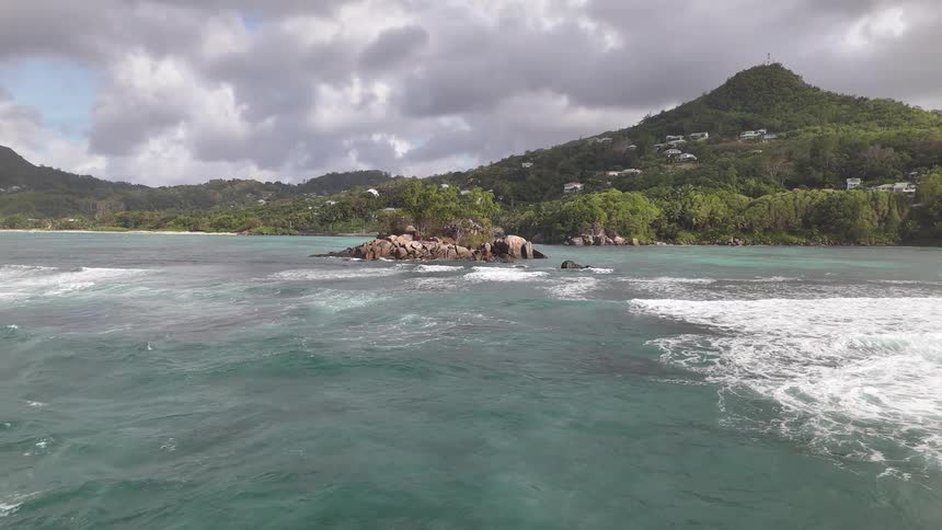 A dramatic aerial shot circles a small rocky islet as large waves and white water crash on all sides with a larger island visible in the background