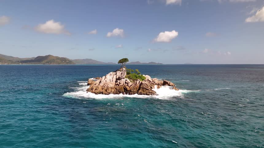 A small rocky island with a single windswept tree rises from turquoise ocean waves under a clear blue sky