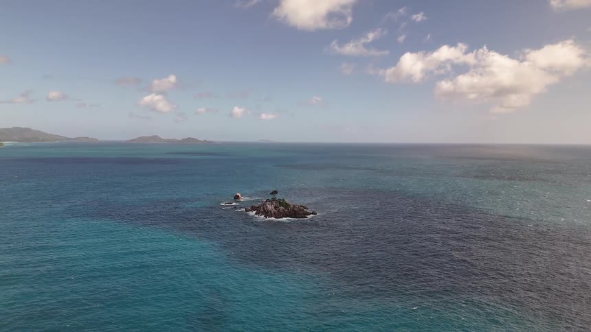 An aerial shot pulls back to reveal a tiny isolated rocky islet with a few trees adrift in a vast and beautiful blue ocean