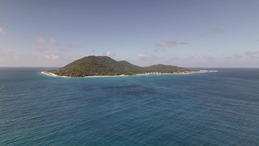 The video shows a breathtaking aerial view of a small lush green island surrounded by vibrant turquoise ocean the sky is clear with a few scattered white clouds enhancing the tropical scenery