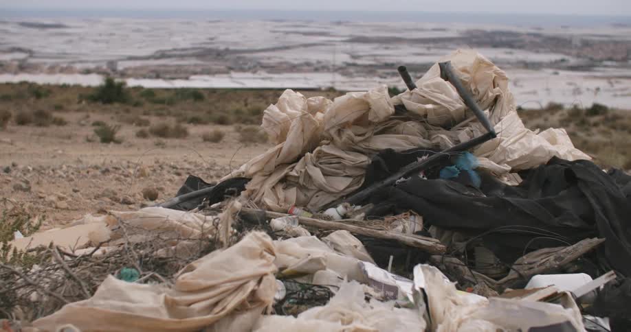 Plastic sheets and debris are strewn across dry terrain near a calm body of water under a hazy sky