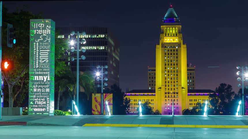 Los Angeles city hall glows in vibrant yellow and purple hues at night framed by urban lights and a multilingual grand park sign