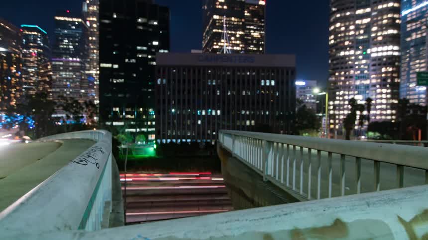Vehicles streak across a city street under a pedestrian bridge tagged with graffiti while lit buildings glow in the nighttime