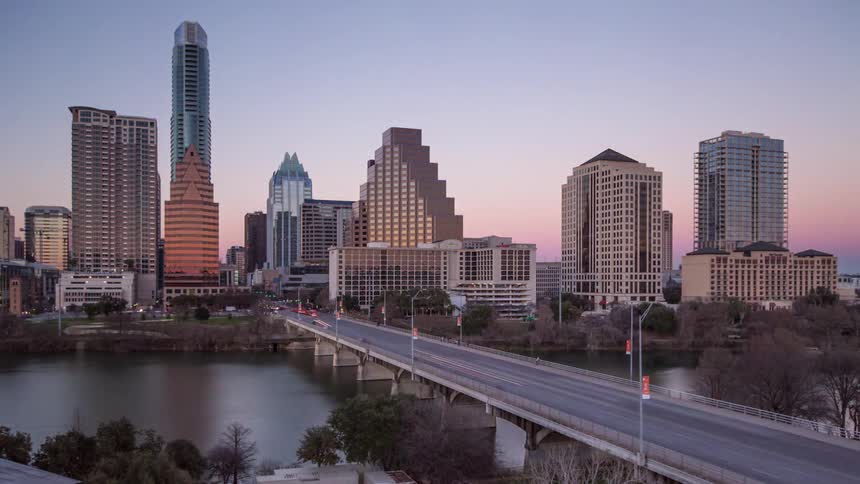 Austin's skyline transitions from daylight to a glowing night scene with building lights reflecting off the colorado river and streaks of car lights trailing across the foreground bridge