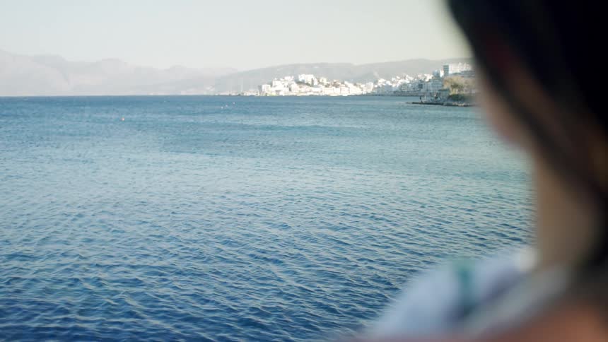 A woman with short dark hair sits wrapped in a green blanket gazing out at a calm sea with distant mountains under overcast light