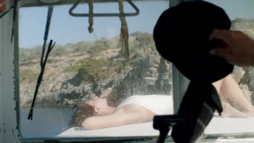 A woman sunbathes on a boat deck near a rocky coastline seen through a cabin window