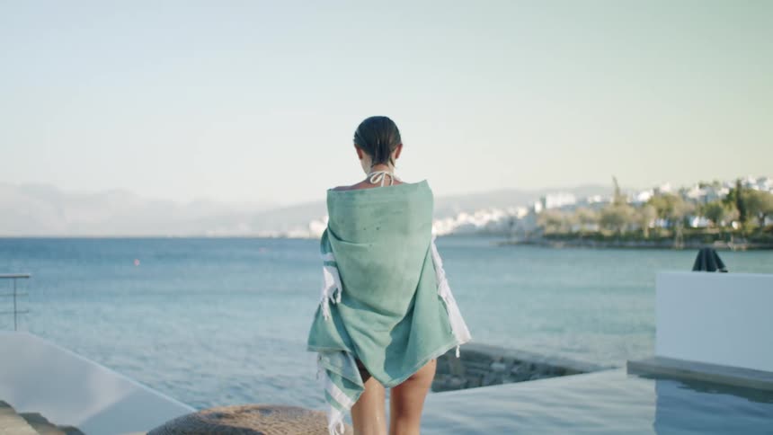 Woman in a swimsuit with wet body laying a towel on the edge of a pool and the sea