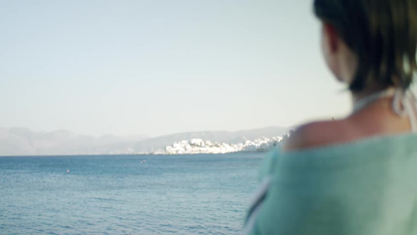 A woman with short dark hair sits wrapped in a green blanket gazing out at a calm sea with distant mountains under overcast light