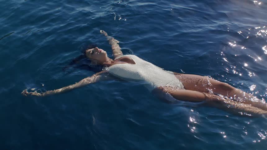A woman in a white swimsuit floats on her back in deep blue ocean water with arms and legs spread wide