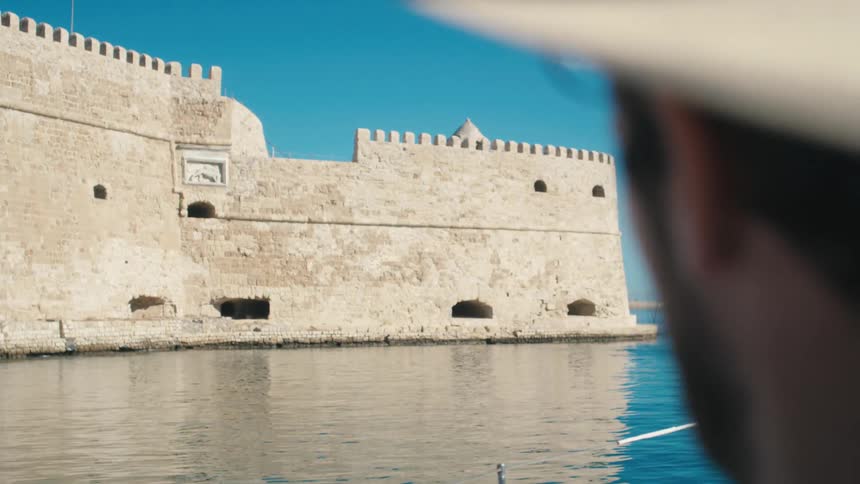 The video shows a historical stone fortress or castle wall situated on the edge of a body of water possibly a sea or large river under a clear blue sky the camera pans slightly revealing more of the ancient structure