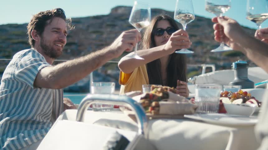 Friends raise glasses in a cheerful toast during a sunny outdoor meal by the water