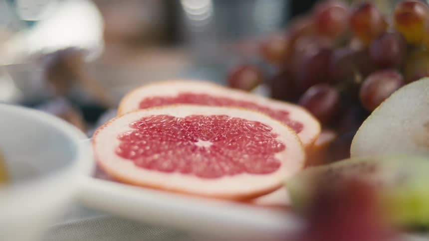A grapefruit sliced in half is featured prominently in the foreground of a closeup shot the image includes blurred grapes and other fruit in the background set against a blurry backdrop with reflections of light