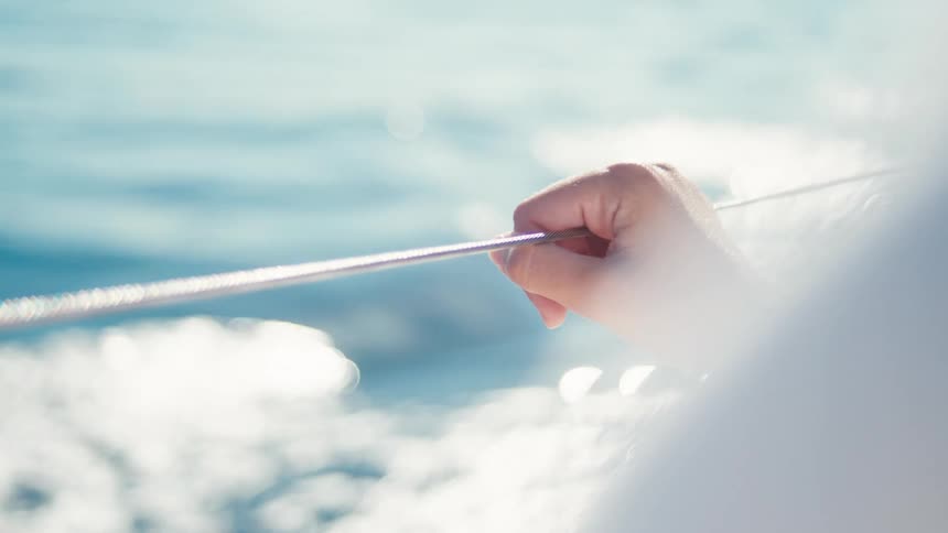 A hand is seen gripping a rope or line on a boat with the blue ocean and sunlight reflecting off the water in the background the focus is on the steady grip and the movement of the boat through the water