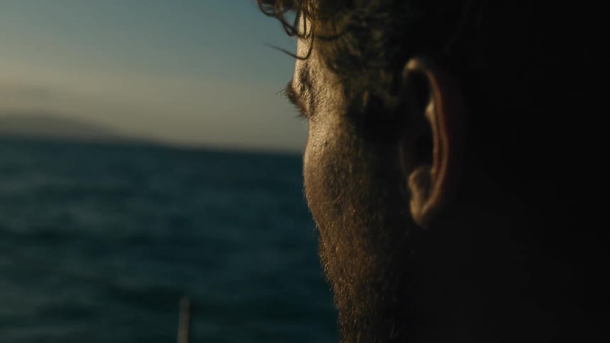 A person is shown in profile looking out at a dark calm body of water possibly at dusk or dawn the scene is characterized by soft warm light on their face contrasting with the dark hues of the water