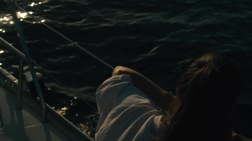 A woman leans on the railing of a boat gazing out at the dark shimmering water in evening light