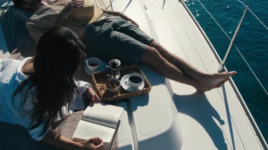 Two people are seen relaxing on the deck of a boat with one person reading a book and the other wearing a straw hat lying down the scene is bright and sunny with the clear blue ocean visible in the background