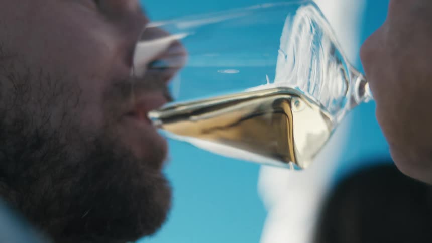 A closeup captures a man sipping white wine from a glass under a bright blue sky