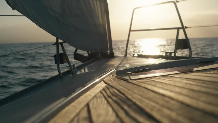 The video shows an eyelevel shot from the deck of a sailboat or yacht looking out at the open ocean the sun is visible on the horizon creating a bright reflection on the water and through the boats rigging