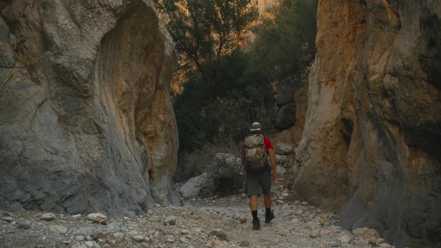 Man with backpack and hat walking along a trail between rocks and mountains