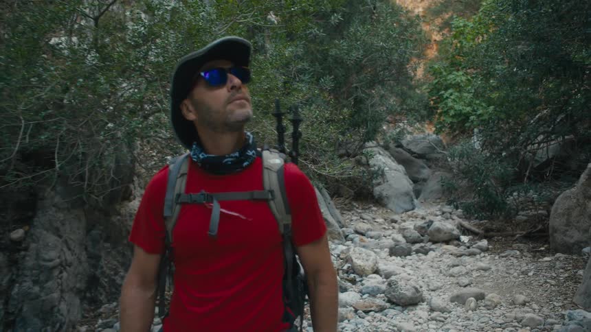 A male hiker wearing a hat and sunglasses with a backpack is walking through a rocky forested area he looks upwards and around seemingly observing his surroundings on a bright day