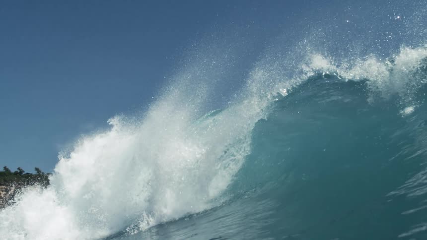 A vibrant blue ocean wave curls and breaks with a powerful spray of white foam against a clear blue sky the crest of the wave is illuminated by sunlight highlighting its transparent qualities before it crashes down