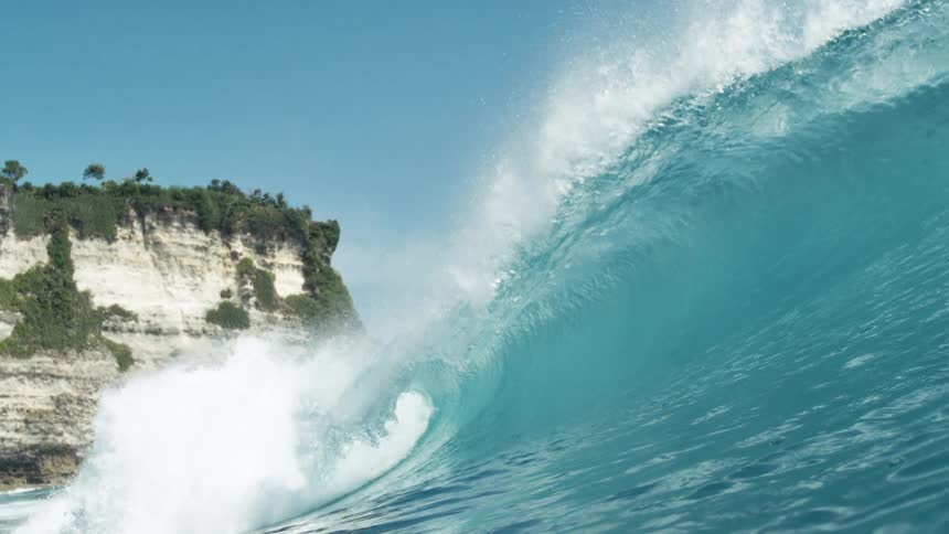 The video shows a powerful blue ocean wave cresting and breaking with white foam and spray visible at the top the sky above is clear and light blue and a distant rocky cliff face is partially visible on the left side of the frame