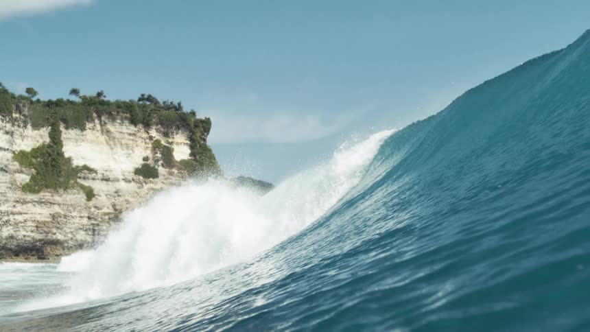 A large ocean wave with white foam crests in the foreground as a distant cliff covered in greenery rises under a clear blue sky