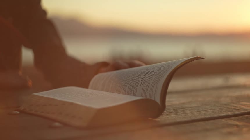 A persons hands are shown turning the pages of an open book resting on a wooden surface the background is blurred with a warm golden light suggesting either sunrise or sunset