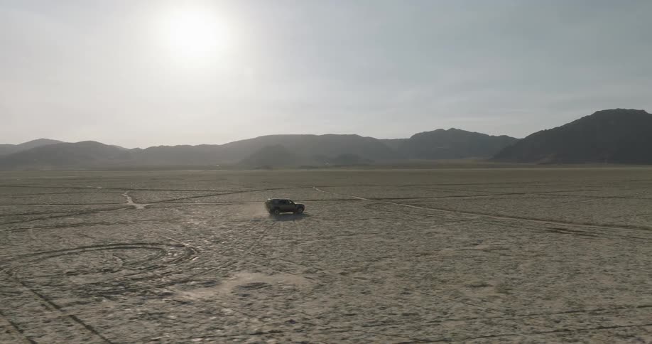A lone vehicle drives across a vast desert plain with dry cracked earth and distant mountains under harsh sunlight