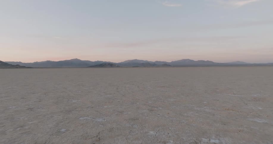 A vast cracked desert stretches toward jagged mountain ranges under a pale cloudless sky