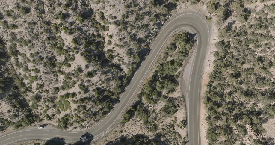 A car drives along a winding asphalt road through a dry rocky desert with sparse shrubs and pale grass