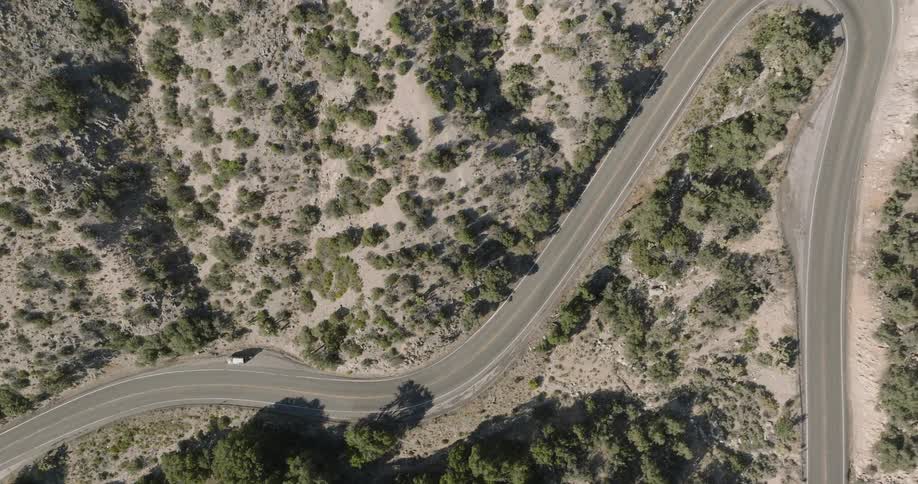 An aerial shot captures a white car driving along a winding road through a mountainous arid landscape sparsely covered with trees the road cuts through the rugged terrain highlighted by the strong sunlight and shadows