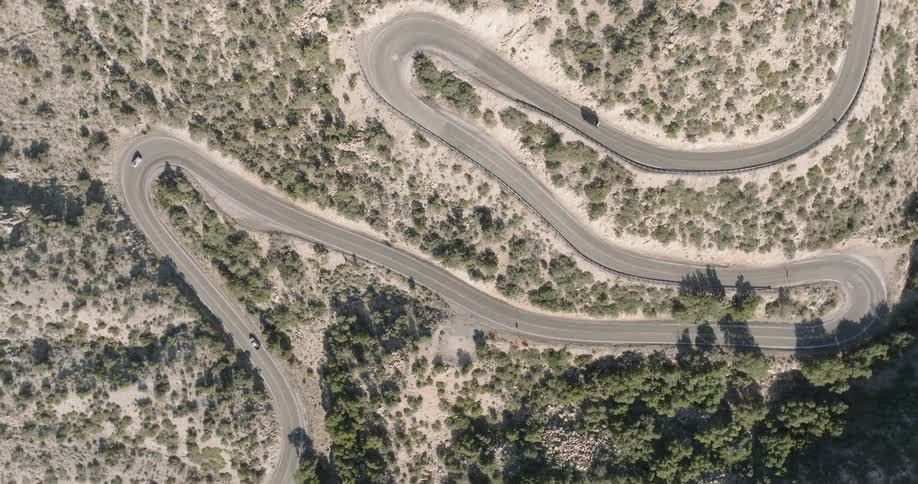 A motorcycle rides along a winding asphalt road through a dry rocky desert with sparse shrubs and pale grass
